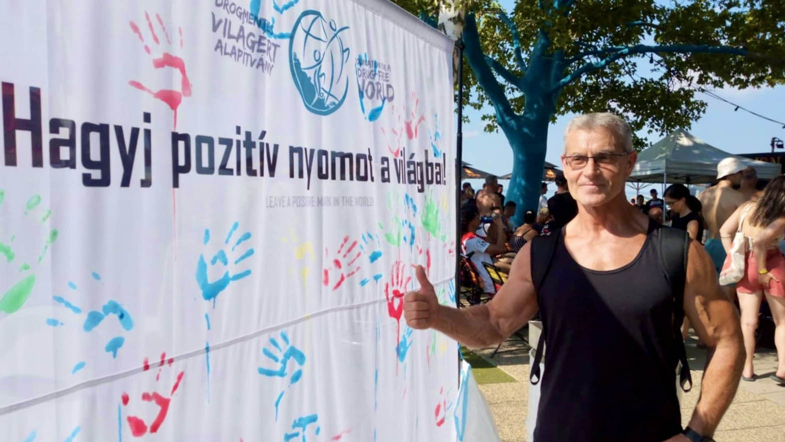 Bodybuilder a siófoki Bodymedia Beach fesztiválon, a Drogmentes Világért Alapítvány ’Hagyj pozitív nyomot a világban!’ molinója előtt, hüvelykujját mutatva